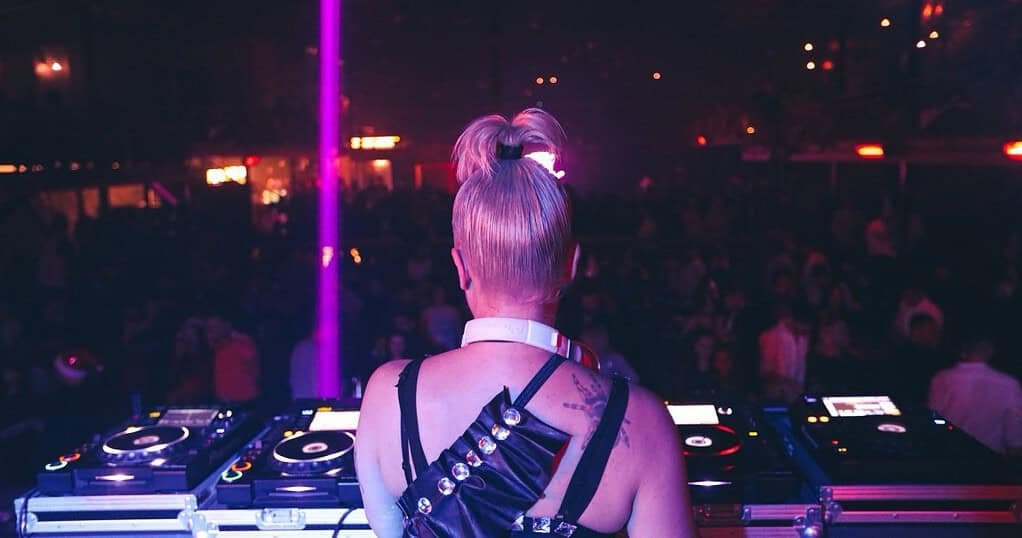 A female techno DJ performing to a crowd under club lights