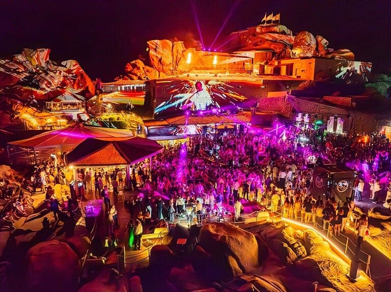 Crowd at Phi Beach Sardinia at night with projections mapped across the fortress rocks